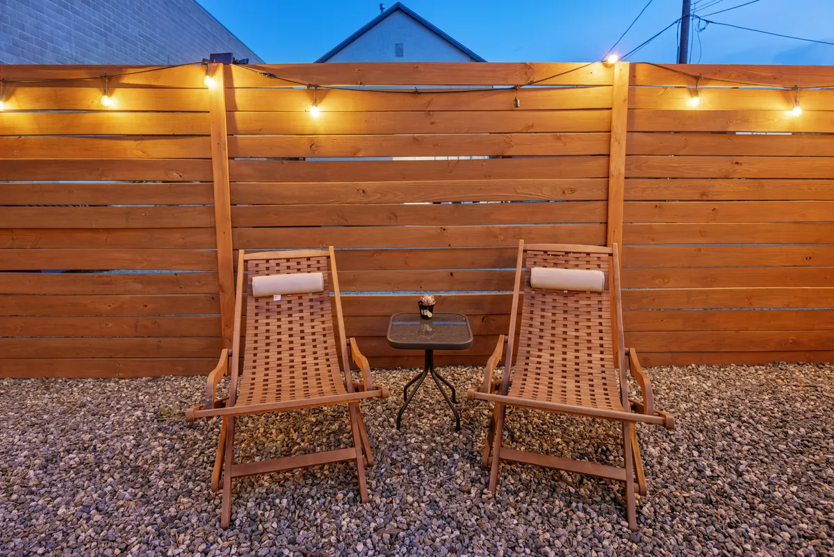 This exterior scene features wooden chairs arranged around a small table under the glow of string lights, creating a cozy and inviting backyard setting perfect for relaxation or casual conversation. The ambiance suggests an ideal space for unwinding after work or taking a peaceful break during the day.