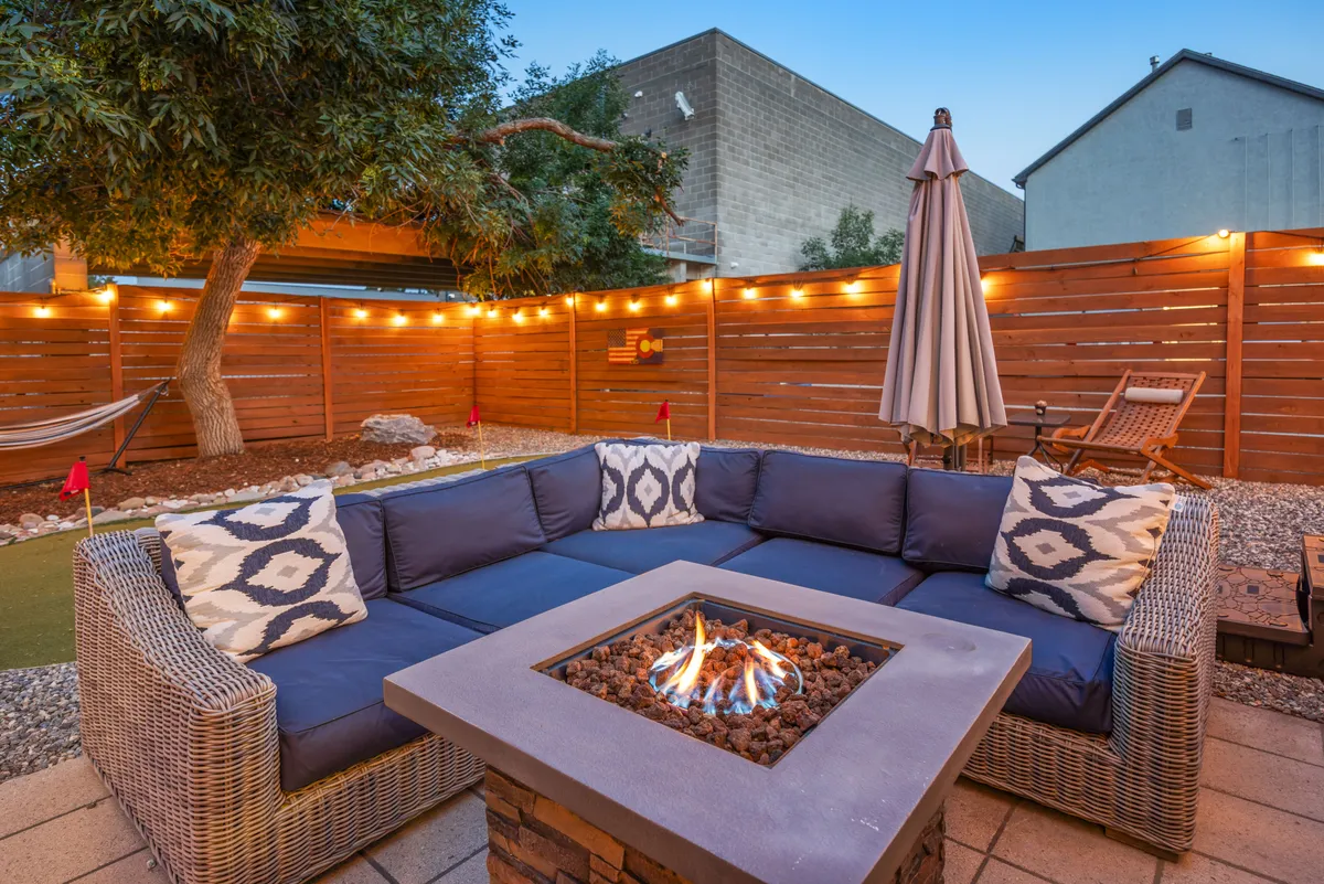 A cozy, well-lit patio area featuring a fire pit, sectional sofa, string lights, an umbrella, and a hammock. The space is arranged for intimate gatherings or relaxation after a day spent exploring city attractions.