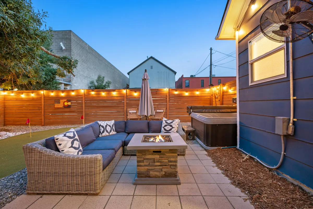 This image showcases the heart of the backyard oasis, featuring a cozy fire pit surrounded by comfortable outdoor seating and a hot tub nearby. String lights hang overhead, casting a warm glow and creating an inviting evening ambiance. The space appears perfect for both social gatherings and relaxed nights under the stars.