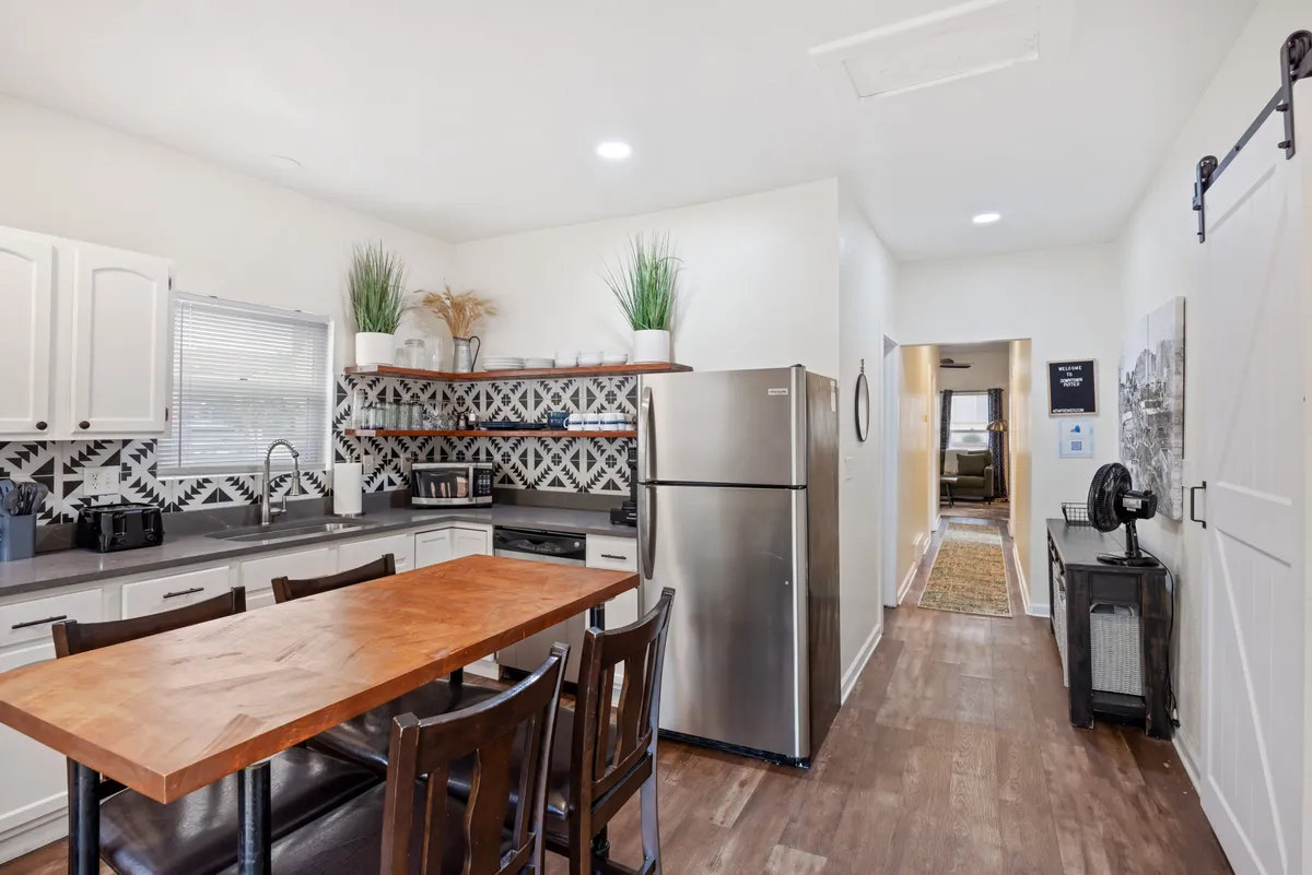 A view of the eat-in kitchen, featuring a wooden dining table, decorative tile backsplash, stainless steel refrigerator, and open shelving adorned with plants. The space is warm and welcoming, ideal for cooking, sharing meals, and casual gatherings.