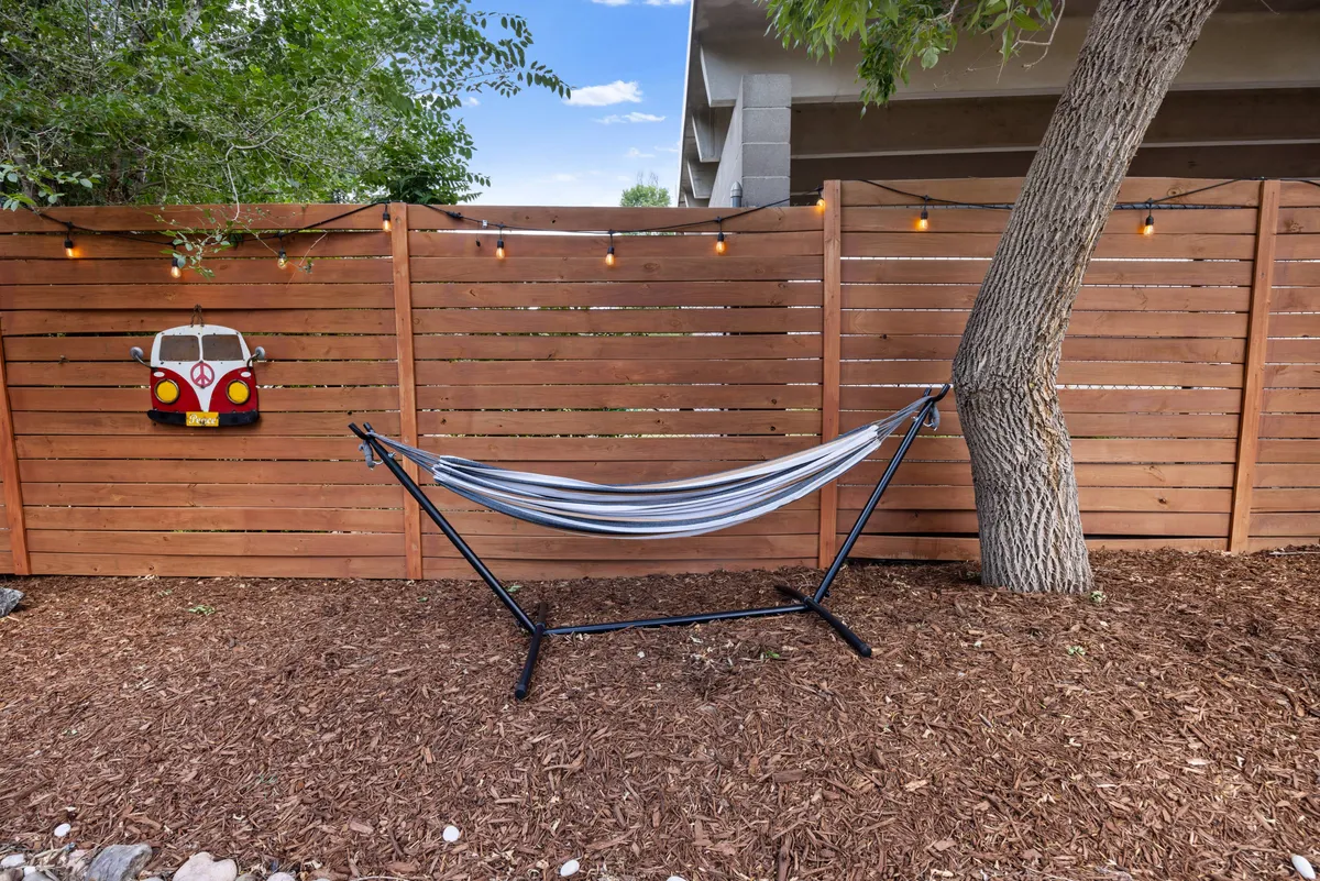 A tranquil corner of the backyard featuring a hammock strung between supports, a decorative van sign, string lights, and a wooden fence. The area invites leisurely lounging, reading, or simply soaking in the outdoor ambiance.