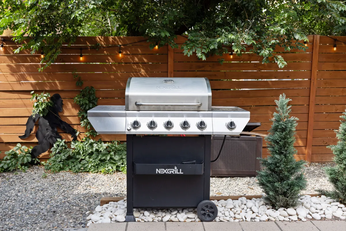 A bright outdoor grilling area featuring a modern grill, surrounded by greenery and framed with a wooden fence. The setup is ideal for barbecues and outdoor feasts, with ample space for cooking and socializing.