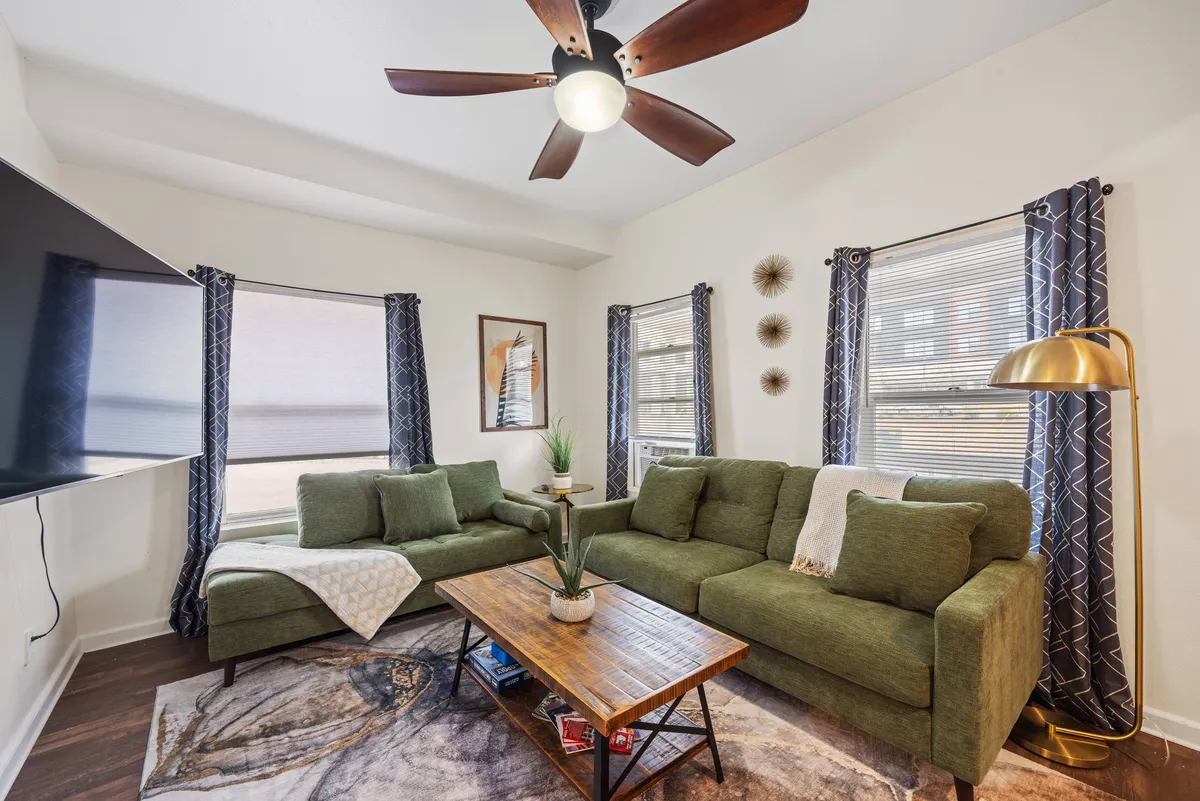 This interior shot centers on a green sectional sofa with chaise, a wooden coffee table, a TV mounted on the wall, decorative wall art, and a floor lamp. The space feels lived-in and designed for comfort and togetherness, inviting guests to relax, watch TV, or enjoy conversation.