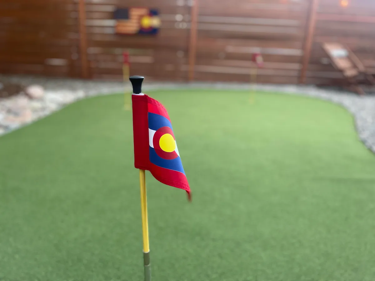 This image showcases a lively backyard area with vibrant artificial turf, golf flags for a putting green, and a wooden fence enclosing the space. The setting is open and inviting, ideal for fun outdoor games and gatherings.