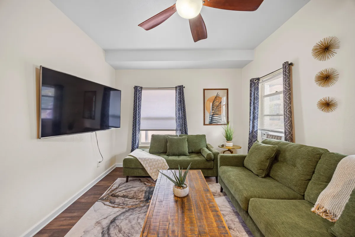 A cozy living room setup with plush green sofas, a large TV, a wooden coffee table, decorative wall art, and a leafy plant in the corner. The space feels inviting for relaxing, reading, or gathering after work. The overall look is contemporary and comfortable, seamlessly extending the workspace atmosphere into a living area.