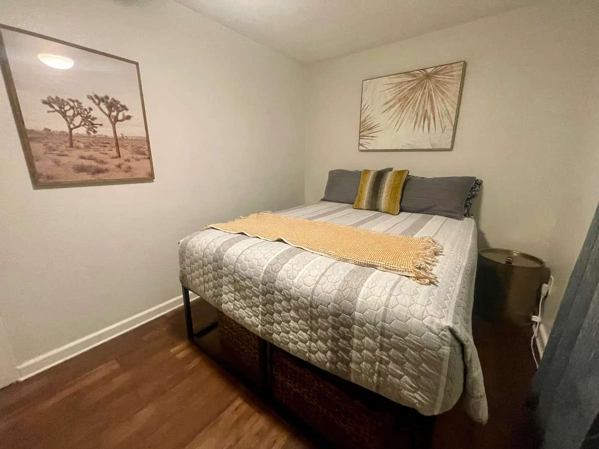 A cozy and serene bedroom with textured bedding, decorative pillows, wall art featuring palm leaves, and a framed photograph of Joshua trees. A window brings in natural light, creating an inviting space for rest and rejuvenation.