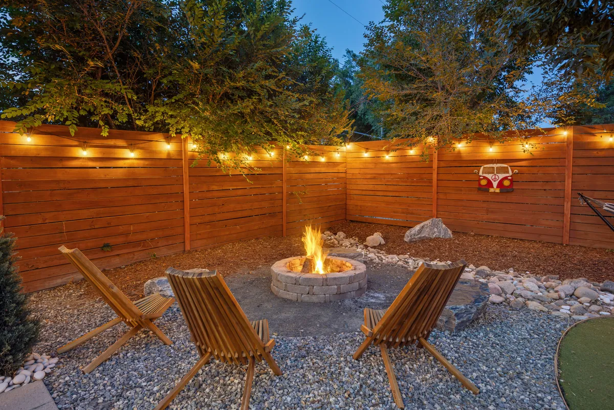 The focus is on a cozy fire pit surrounded by wooden chairs, all beneath twinkling string lights. The scene suggests warmth and social connection, ideal for evening relaxation, storytelling, or making s’mores.