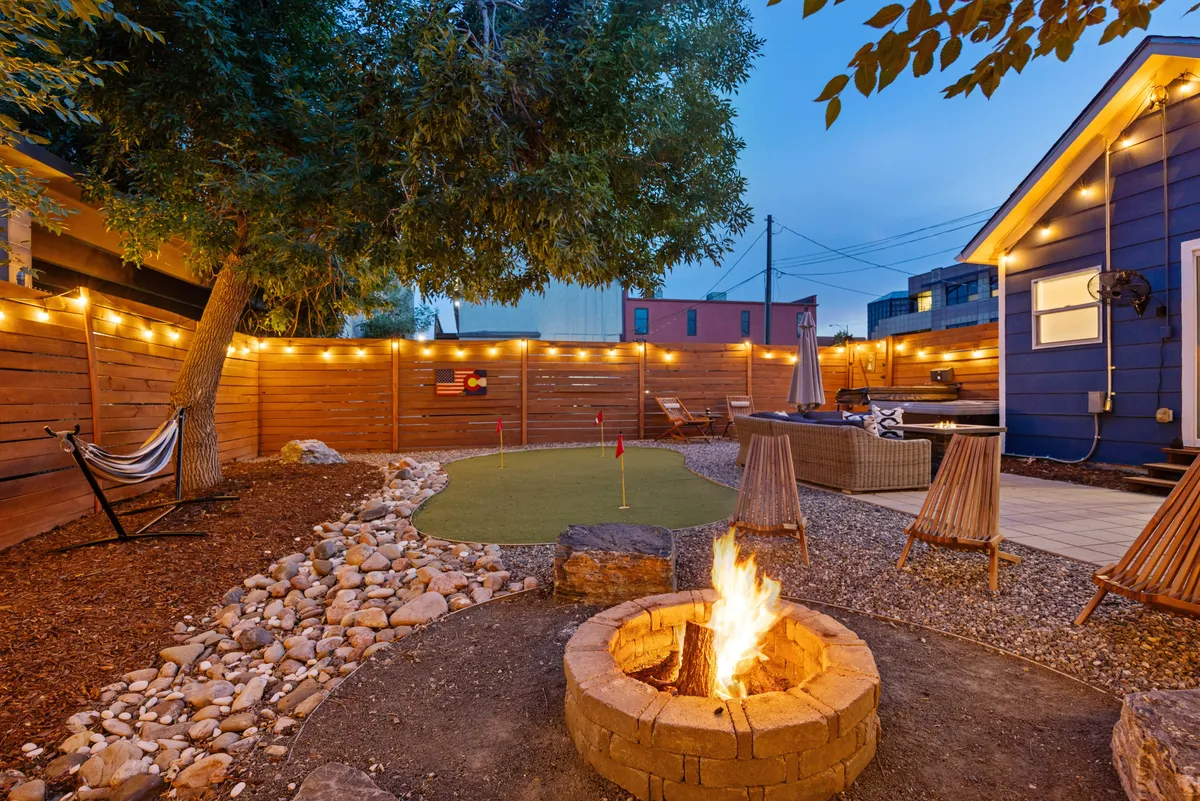 A vibrant backyard scene with a fire pit as the focal point, complemented by a lush putting green, a hammock for lounging, inviting seating areas, and twinkling string lights. The layout suggests multiple zones for different activities, from practicing golf to enjoying quiet moments in the hammock.
