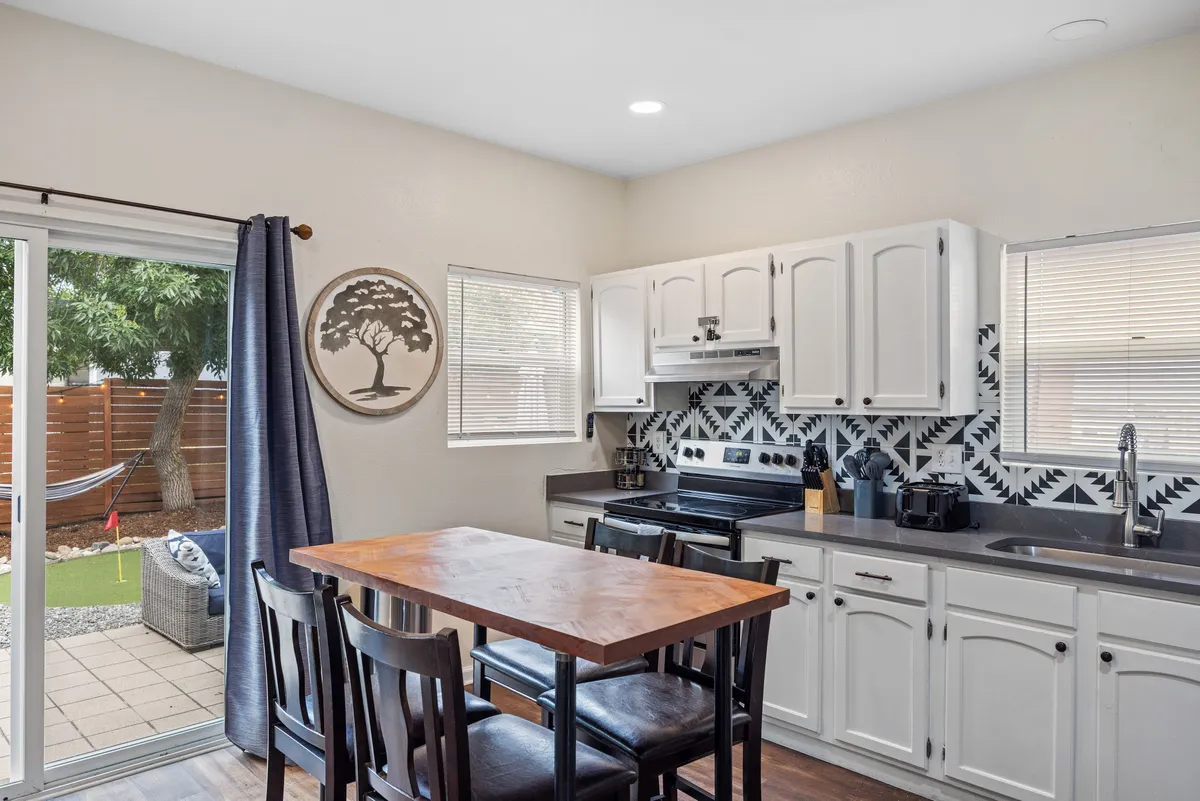 A cozy dining area features a wooden table set by a window with light streaming in, complemented by a black and white patterned backsplash and modern kitchen appliances. The inviting scene hints at relaxed breakfasts or coffee mornings, creating a warm atmosphere for starting the day together.